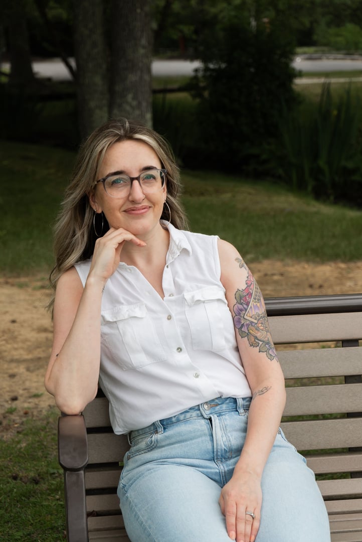 Emilee, a white woman with glasses, tattoos, and brown and blonde hair, is wearing a white button-down tank top and jeans. She's sitting on a park bench and resting her chin on her hand. 