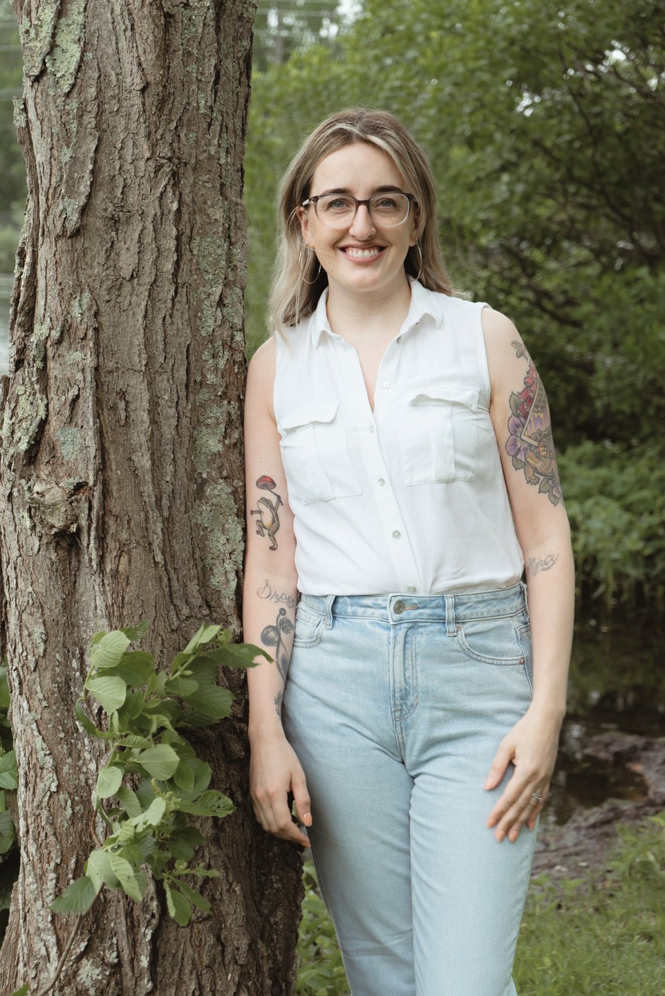 Emilee, a white woman with brown and blonde hair, is wearing a white button-down tank top and jeans, leaning against a tree. She has tattoos and wears glasses. Emilee is a virtual assistant and this photo is supposed to be friendly and inviting.