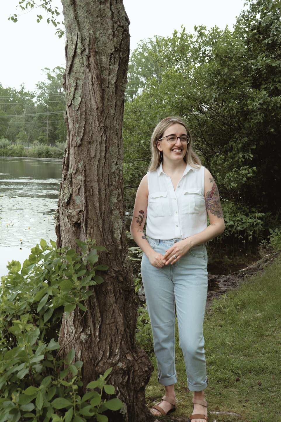 Emilee, a white woman wearing a white button-down tank top and jeans, stands a little awkwardly next to a tree. She really tried to pose well. 